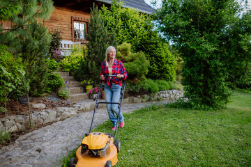 Elderly woman mowing grass with lawn mower in the garden, garden work concept.