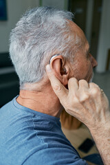 Obraz premium Senior man tunes his hearing aid behind the ear by pressing his finger on setting button. Modern hearing aid, hearing solutions