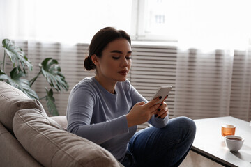 Portrait of young brunette woman with concentrated facial expression sitting on the couch typing on the phone. Joyful female having a video call. Background, copy space, close up.