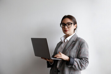 Portrait of a young businesswoman wearing smart casual attire standing with a laptop. Studio shot of a brunette female in eyeglasses posing near white wall. Close up, copy space, background.