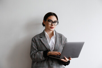 Portrait of a young businesswoman wearing smart casual attire standing with a laptop. Studio shot of a brunette female in eyeglasses posing near white wall. Close up, copy space, background.