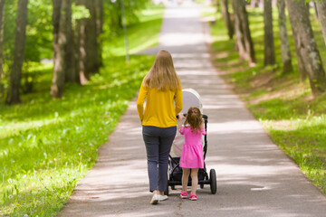 Young adult mother and little daughter pushing white baby stroller and walking at town park in warm...