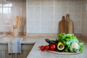 Close up shot of different raw fruits, vegetables and greens, wooden chopping boards on the marble countertop of kitchen with classic style interior. Pastel color facing tiles. Copy space, background.