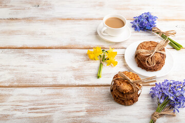 Oatmeal cookies with spring snowdrop flowers bluebells, narcissus and cup of coffee on white wooden background. side view, copy space.