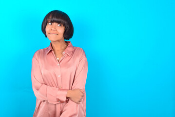 young brunette woman wearing pink silk shirt over blue background with thoughtful expression, looks away keeps hands down bitting his lip thinks about something pleasant.