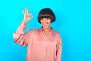 Glad attractive young brunette woman wearing pink silk shirt over blue background shows ok sign with hand as expresses approval, has cheerful expression, being optimistic.