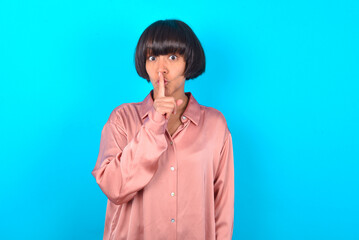 young brunette woman wearing pink silk shirt over blue background makes hush gesture, asks be...