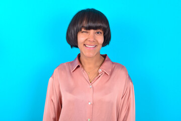Fototapeta premium young brunette woman wearing pink silk shirt over blue background blinking eyes with pleasure having happy expression. Facial expressions and people emotions concept.