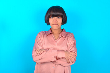 young brunette woman wearing pink silk shirt over blue background bitting his mouth and looking worried and scared crossing arms, worry and doubt.