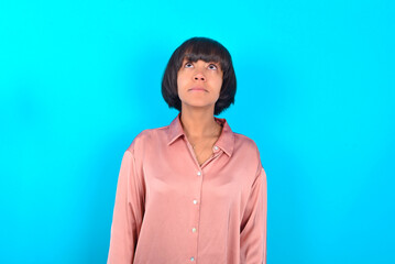 Fototapeta premium young brunette woman wearing pink silk shirt over blue background looking up as he sees something strange.