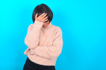young brunette woman wearing pink knitted sweater over blue background making facepalm gesture...