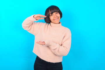 young brunette woman wearing pink knitted sweater over blue background gesturing with hands showing...