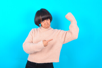 Smiling young brunette woman wearing pink knitted sweater over blue background raises hand to show...