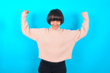 Waist up shot of young brunette woman wearing pink knitted sweater raises arms to show muscles...