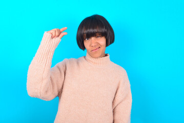 Upset young brunette woman wearing pink knitted sweater shirt over blue background shapes little gesture with hand demonstrates something very tiny small size. Not very much