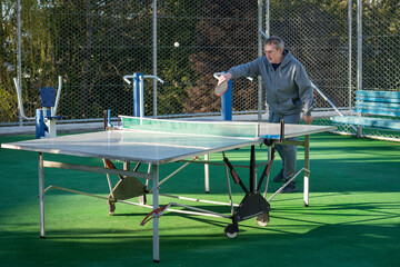 An elderly, gray-haired retired man plays table tennis ping pong on an outdoor sports field on a sunny day. Motion blur on the ball. The concept of an active lifestyle of elderly pensioners