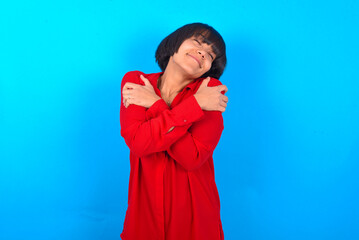 young brunette woman with short hair wearing red shirt over blue background. Hugging oneself happy and positive, smiling confident. Self love and self care.