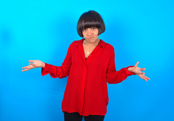 young brunette woman with short hair wearing red shirt over blue background looks uncertain shrugs shoulders.