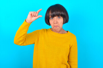young brunette woman with short hair wearing yellow knitted sweater over blue background purses lip and gestures with hand, shows something very little.