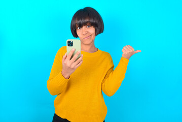 young brunette woman with short hair wearing yellow knitted sweater over blue background  points thumb away and shows blank space aside, holds mobile phone for sending text messages.