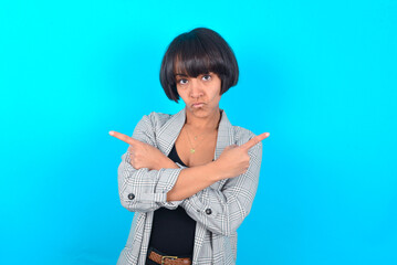 Serious young businesswoman with bob haircut wearing blazer against blue wall crosses hands and points at different sides hesitates between two items. Hard decision concept