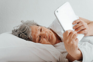 Senior woman using tablet while lying in bed in bedroom. Close-up of sleepy elderly woman reading online or browsing internet while relaxing in room