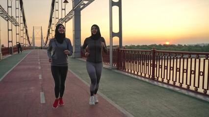 Sporty muslim women in hijabs and sportswear running across urban bridge over river at sunset. Fit arab joggers training together in the evenings, active hobby, healthy sports lifestyle concept