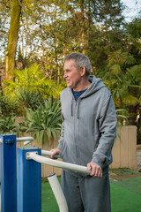 An elderly gray-haired man pensioner in a gray suit is training on simulators on a sports field on the street. The concept of an active healthy lifestyle of elderly pensioners.