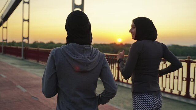 Rear view of sporty muslim females in hijabs and sportswear running across city footbridge at sunset. Healthy arab women enjoying joint cardio workout outdoors in the evenings