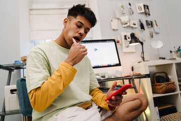 Brunette teenage boy brushing his teeth and using cellphone in bedroom