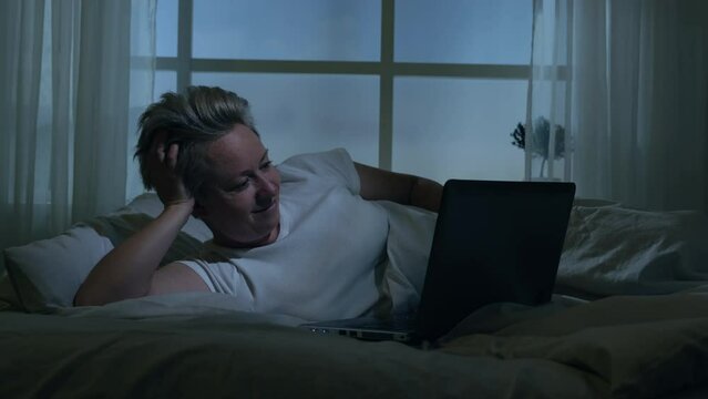 Woman watching video on laptop in bed late at night, movie screening before going to sleep.