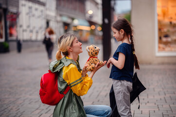 Young blond woman in raincoat and backpack give present toy to little preschooler girl on street