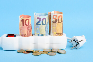 Euro bills in sockets of power strip and plug with coins on blue background. Concept of expensive electricity costs in Europe and rise in energy bill prices.