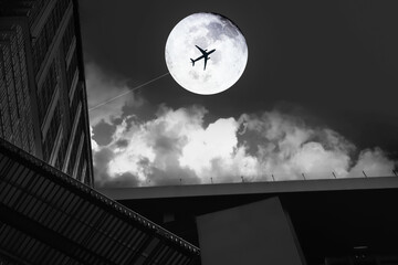 Black and white image of full moon with aircraft silhouette in dark sky background with bright clouds and modern building at night time for travel and transportation background.