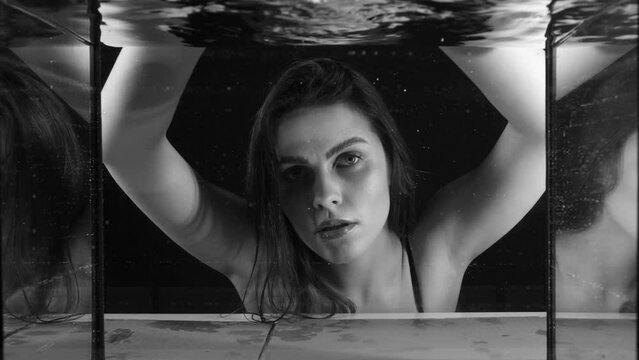 Beautiful young woman with coral lips looking at camera through glass with water drops. Black and white video