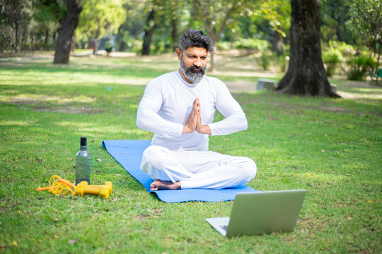 Beard Indian Man Doing Yoga Meditation Exercise In The Park While Using Laptop For Online Class Or Virtual Tutorials. Fitness And Healthy Lifestyle.