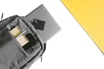 Obraz premium Gray urban backpack with a laptop, notepad, pen, external ssd and smartphone on white and yellow background. Lifestyle concept with gadgets. Backpack concept for the modern student.