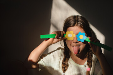 Child girl closes her eyes with paper yellow blue flowers and shows a funny tongue in the rays of the sun, indoors, Ukrainian child © Julia