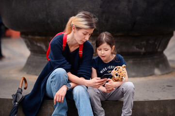 Young woman in spring outfit sits with little preschooler girl on stairs of old city fountain