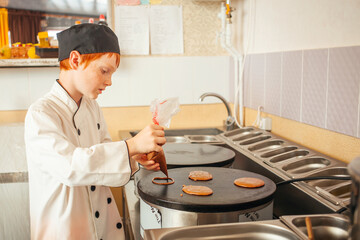 boy cooks pancakes on an industrial professional pancake maker, kitchen in cafe, little chef. Pours dough on stove.