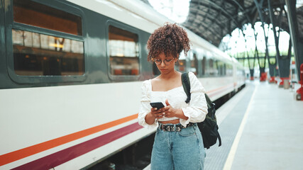 Close-up portrait of a young woman in glasses with a smartphone and backpack stands at the station. Positive woman using mobile phone outdoors in urban background.