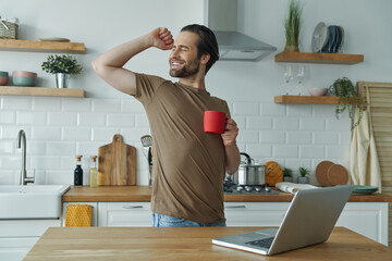 Handsome young man smiling and gesturing while enjoying coffee at the kitchen