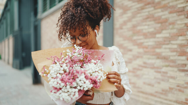 Young Woman In Glasses Walks With A Bouquet Of Flowers Along The Street Of The Old City And Listens To Music In Wireless Headphones