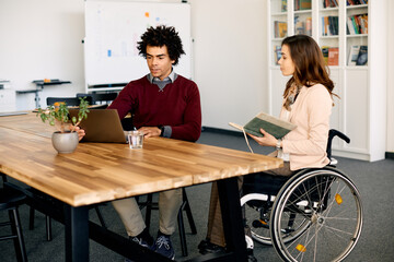 African American businessman and his colleague in wheelchair cooperate while working on laptop in...