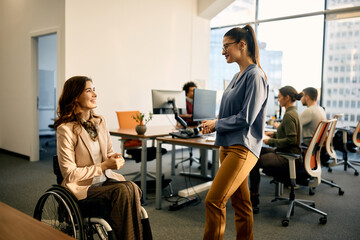 Happy businesswoman in wheelchair talks to female colleague in the office.