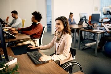 Happy businesswoman in wheelchair works on desktop PC at IT office.
