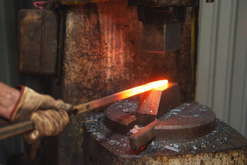 Professional male blacksmith forming red hot metal on an anvil in interior blacksmith workshop. - obrazy, fototapety, plakaty