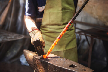 Professional male blacksmith forming red hot metal on an anvil in interior blacksmith workshop.