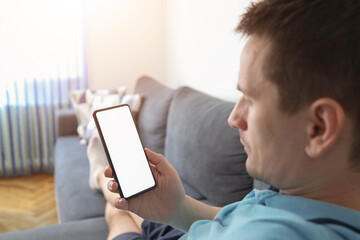 View of man hand holding smart phone with blank copy space screen for your text message or information content. Reading text message on cell telephone at living room.