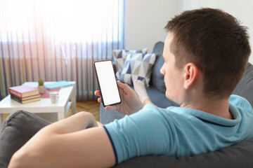 Young man user customer hold smart phone mock up white screen in hand use mobile shopping app, check social media news, texting mobile sms order food delivery sit on sofa at home. Over shoulder view